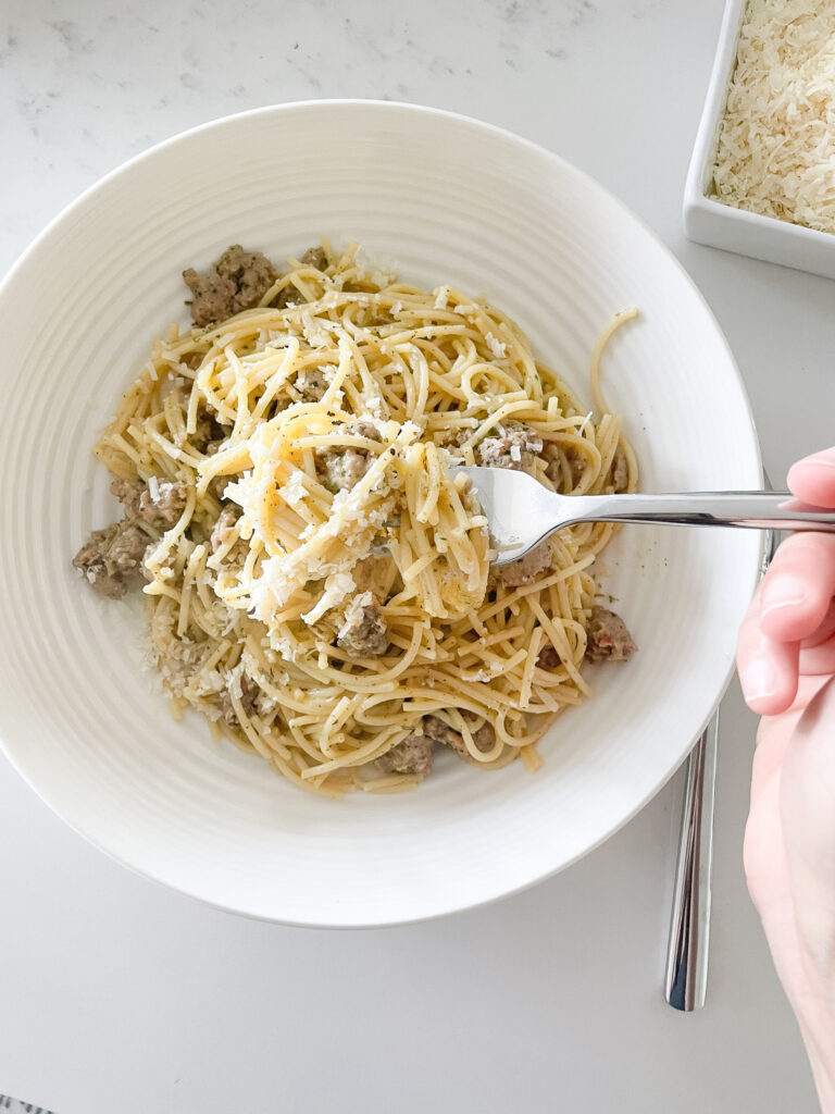 Sausage carbonara on a fork with the bowl of pasta in the background and a bowl of parmesan cheese.