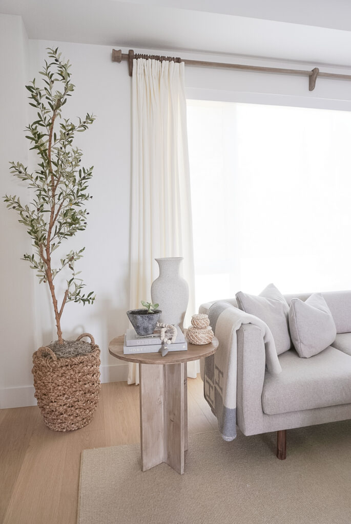 Alward side table from studio mcgee.. excep its a DIY version. It is a pine wood table with an "x" lengthwise for the legs and a round top. It is decorated with a vase, two books, a plant and a set of wooden beads. An olive tree is in the back ground. and a grey sofa next to it.