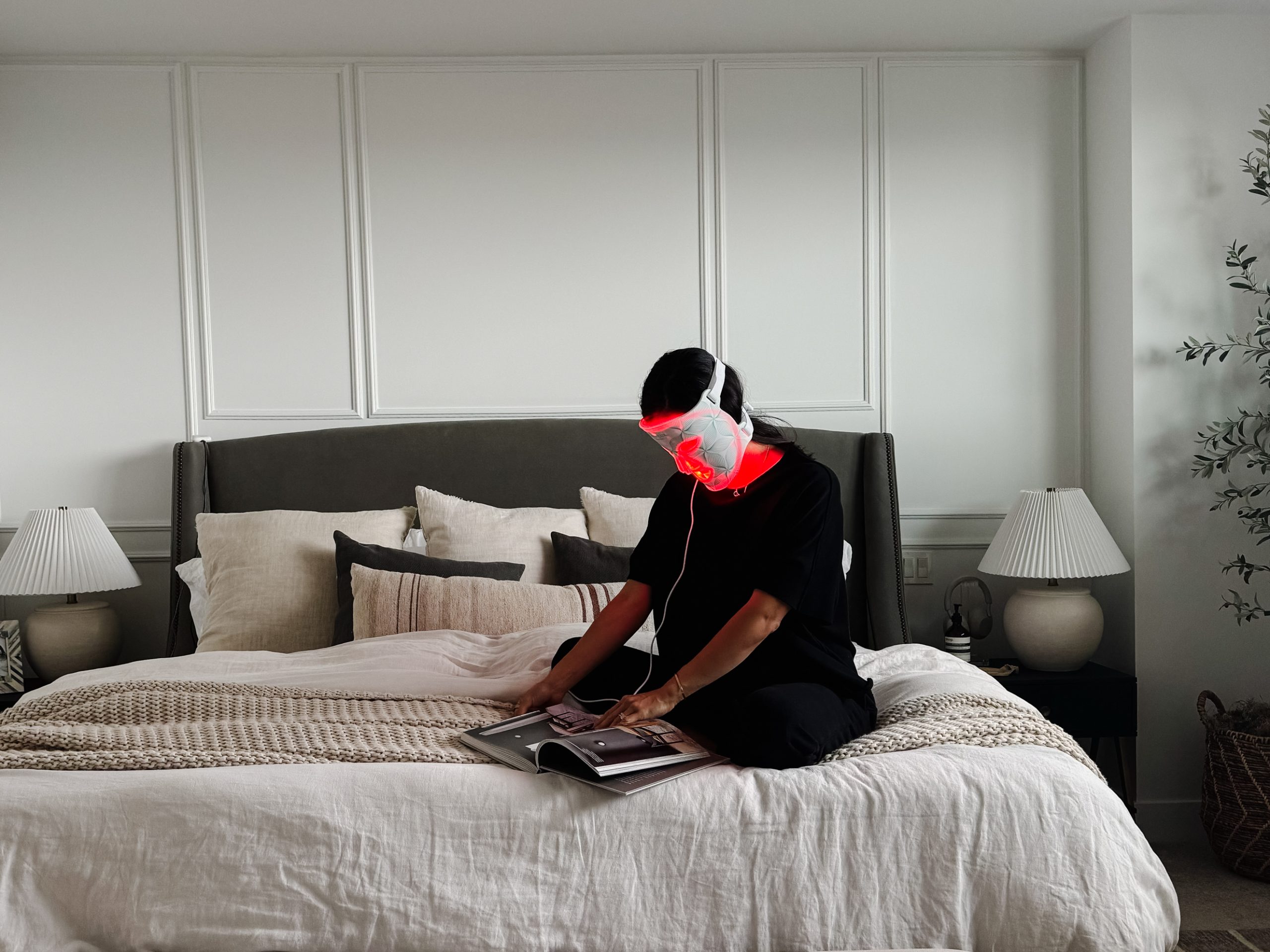 A woman sitting on a bed reading a book wearing a Omnilux Red light therapy contour mask