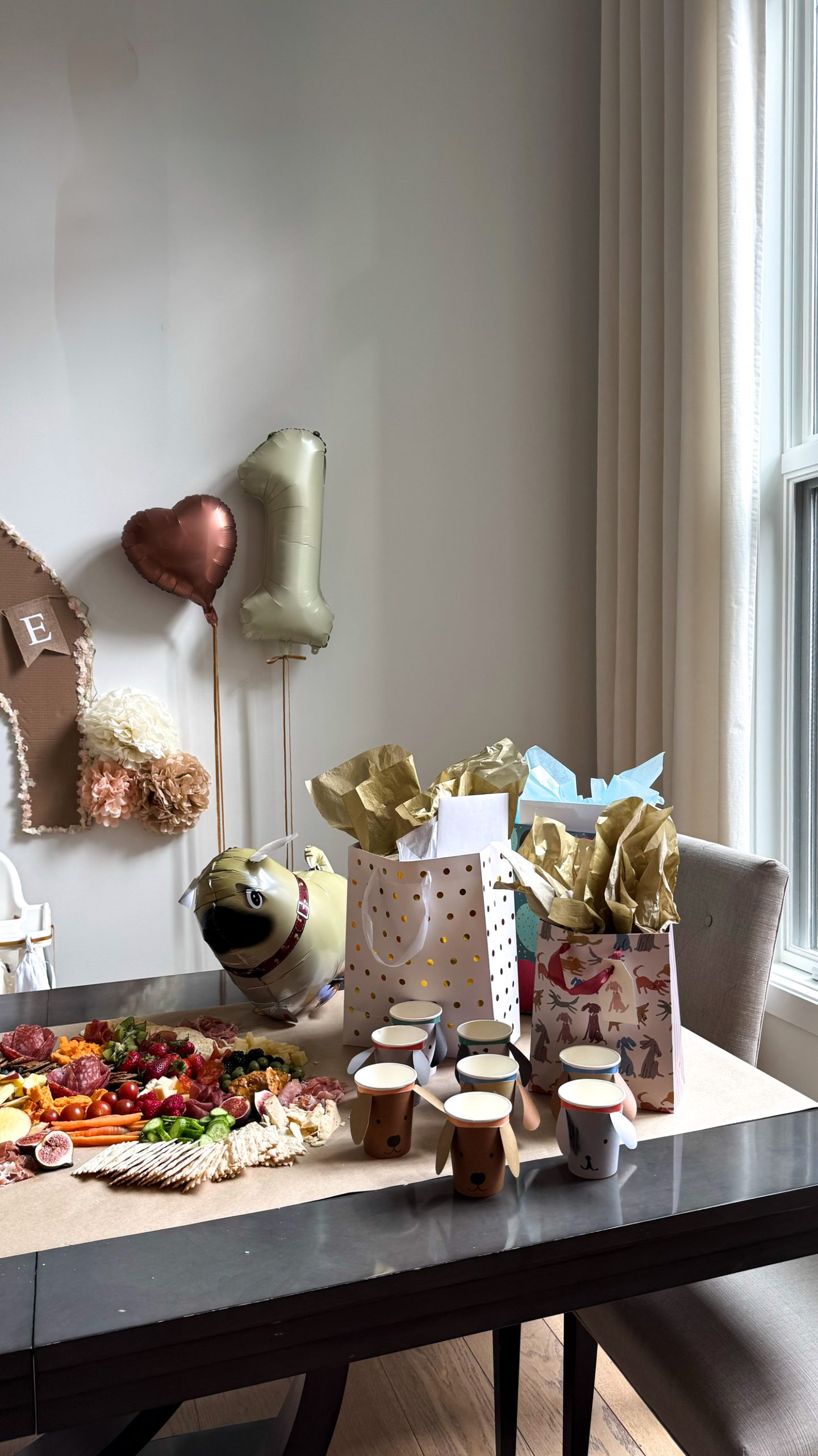 Dog themed first birthday set up with a charcuterie boar don the table, dog blow up balloons, a cardboard dog house, and balloons on the wall.