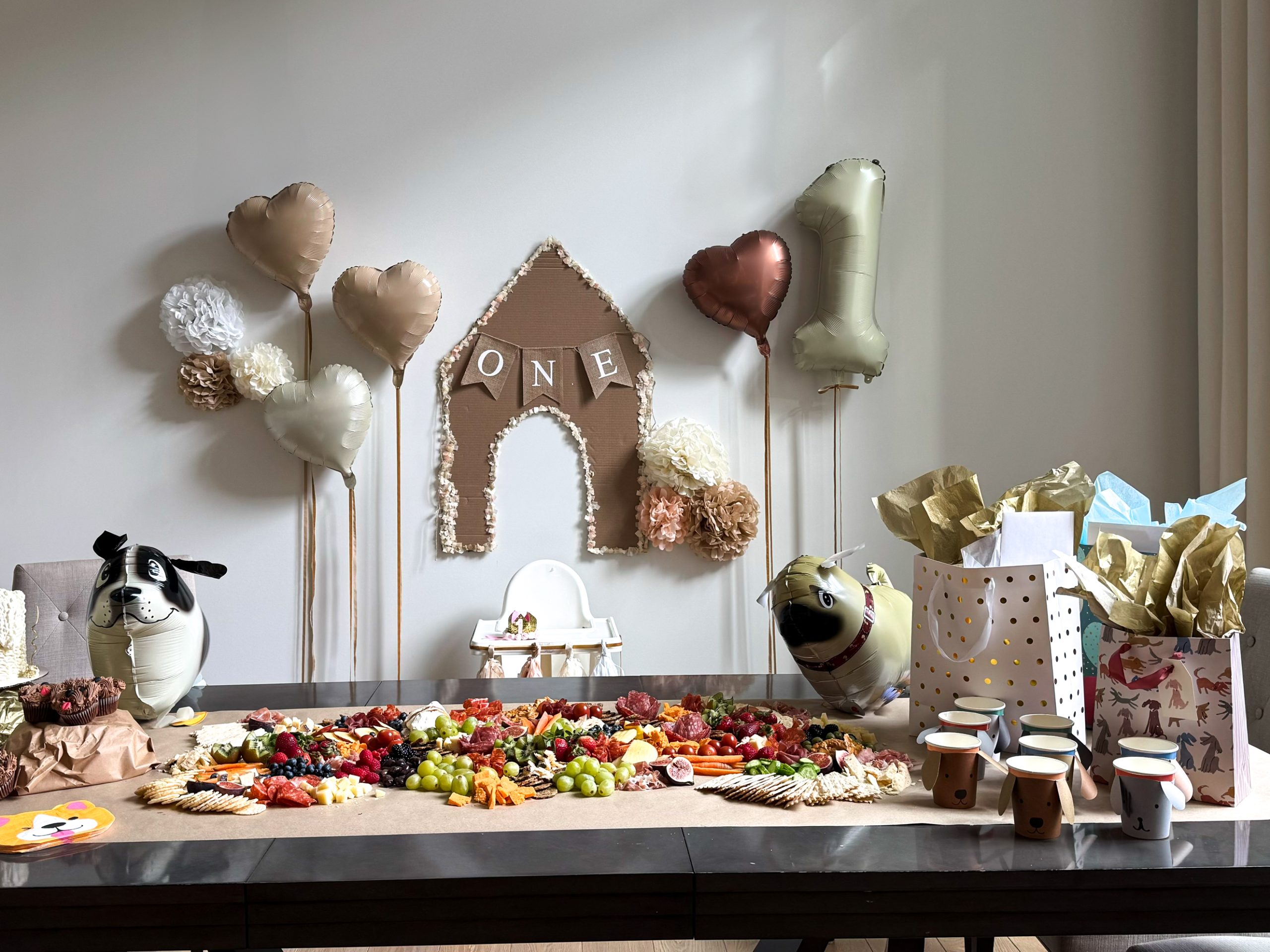 Dog themed first birthday set up with a charcuterie boar don the table, dog blow up balloons, a cardboard dog house, and balloons on the wall.