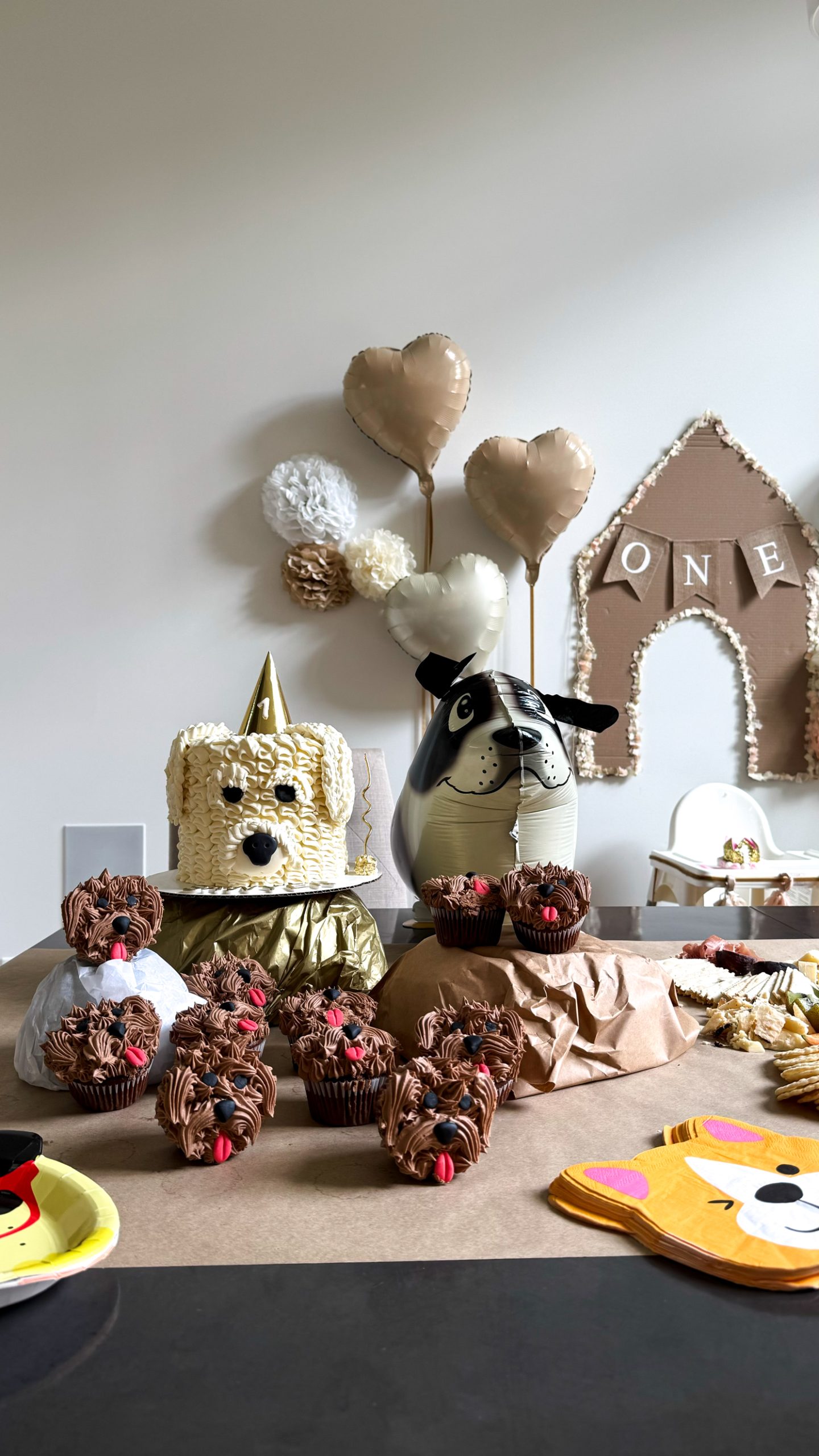 Dog themed first birthday set up with a charcuterie boar don the table, dog blow up balloons, a cardboard dog house, and balloons on the wall.
